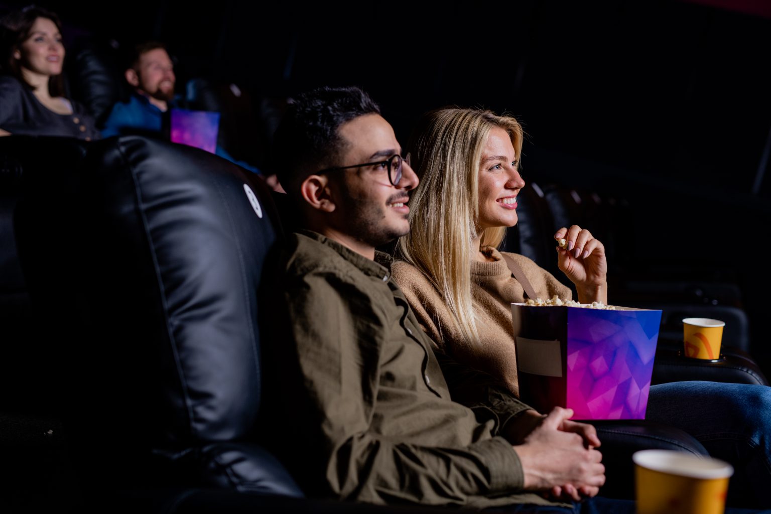 Happy young amorous couple relaxing in cinema in front of large screen while enjoying love story movie